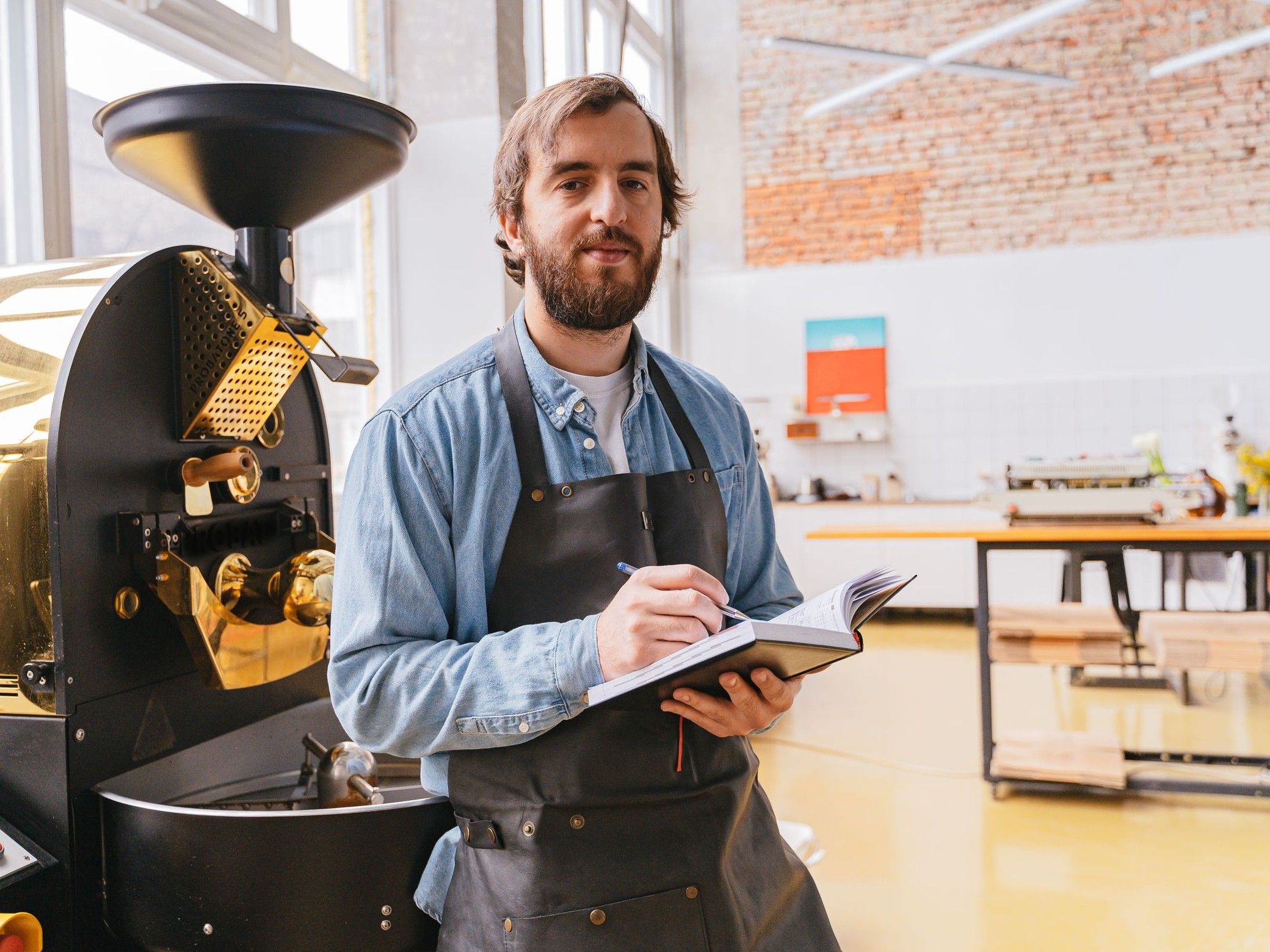 A coffee roastery owner standing by his roaster with a notebook, looking at the camera